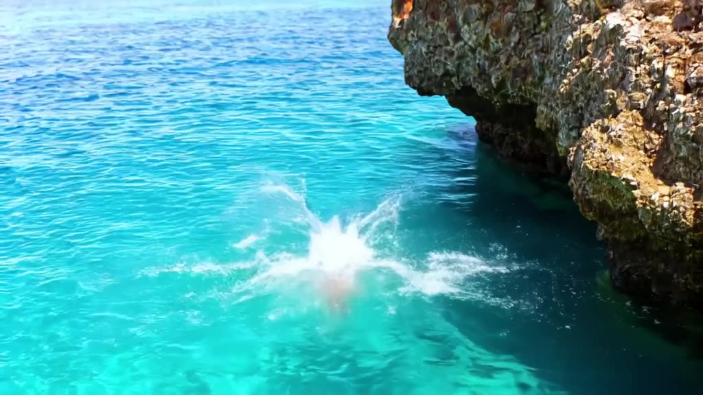A distinguished man with dark shades dives into a crystal-clear blue lagoon from a rocky …