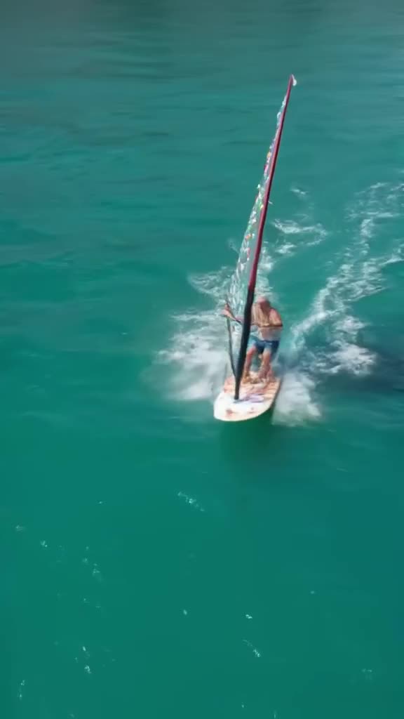 An elderly man windsurfing across a turquoise lake, the sail patterned with confetti. Wat…