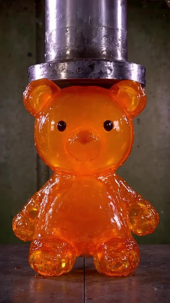 A massive hydraulic press descends slowly onto a giant, translucent orange gummy bear tha…