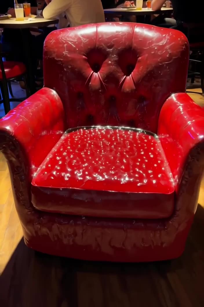 A cozy armchair upholstered entirely in wobbly, translucent red jelly sits in a dimly lit…