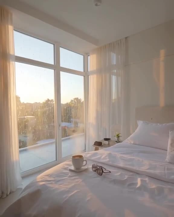 Minimalist white bedroom with floor-to-ceiling windows, rain patters against glass while …