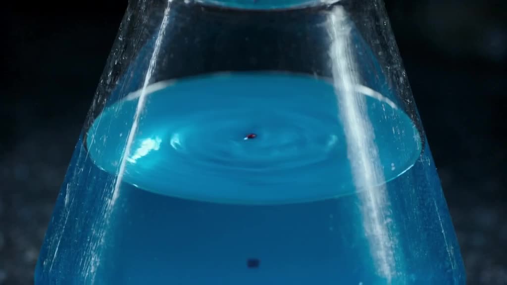 Close up of a pristine beaker filled with bright blue liquid. A single drop of red dye fa…