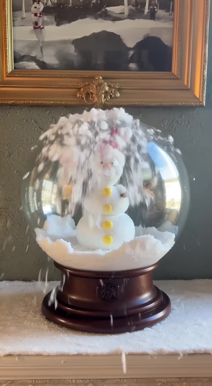 A snow globe sits on a mantel. Inside, a snowman celebrates. The globe shakes, and snow s…