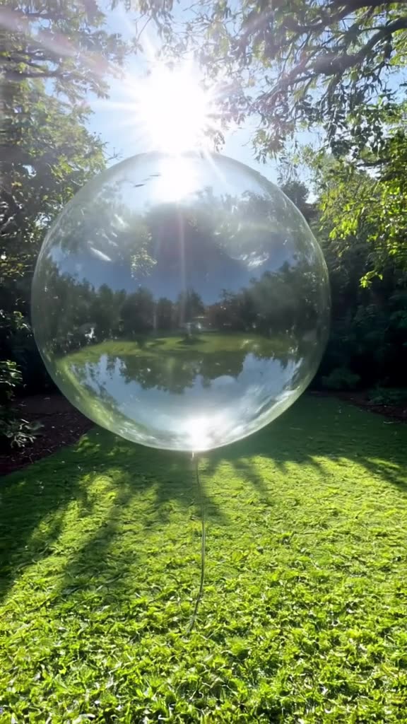 A giant bubble wand blows a massive bubble in a garden. The bubble reflects a miniature l…