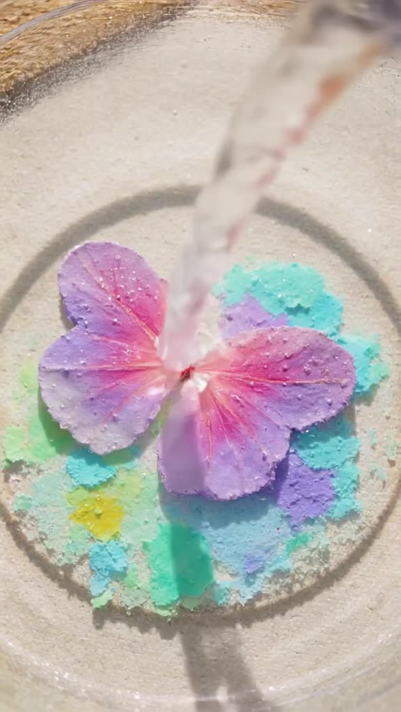 Watercolor paints swirl in a glass bowl, forming a delicate butterfly. The butterfly burs…