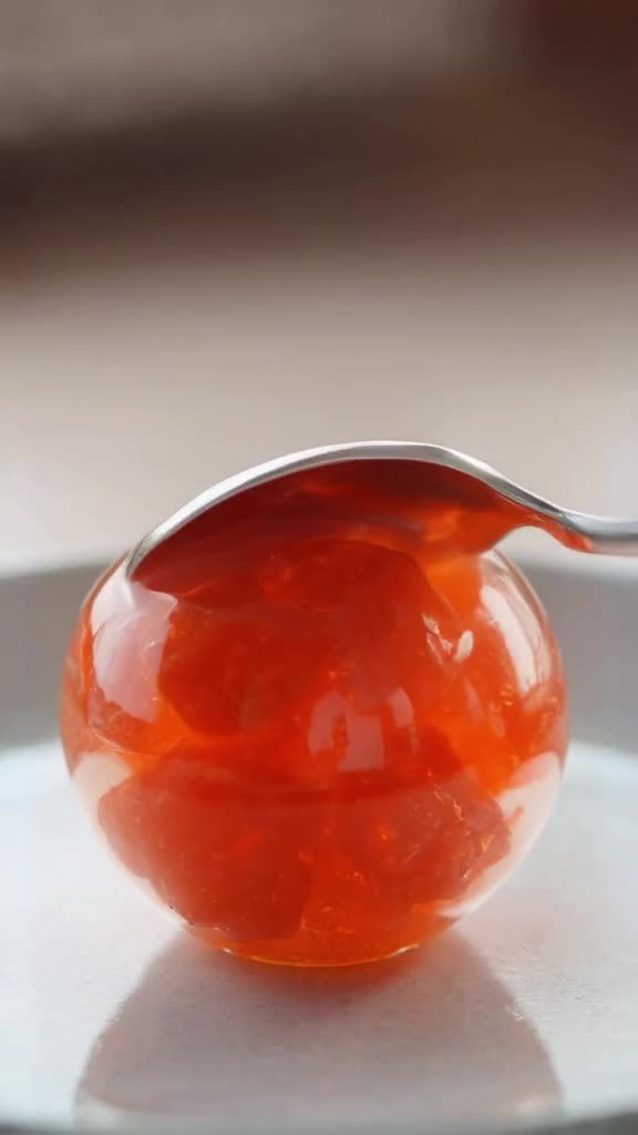 A perfectly spherical gelatin dessert wobbles on a plate. It is clear with suspended oran…
