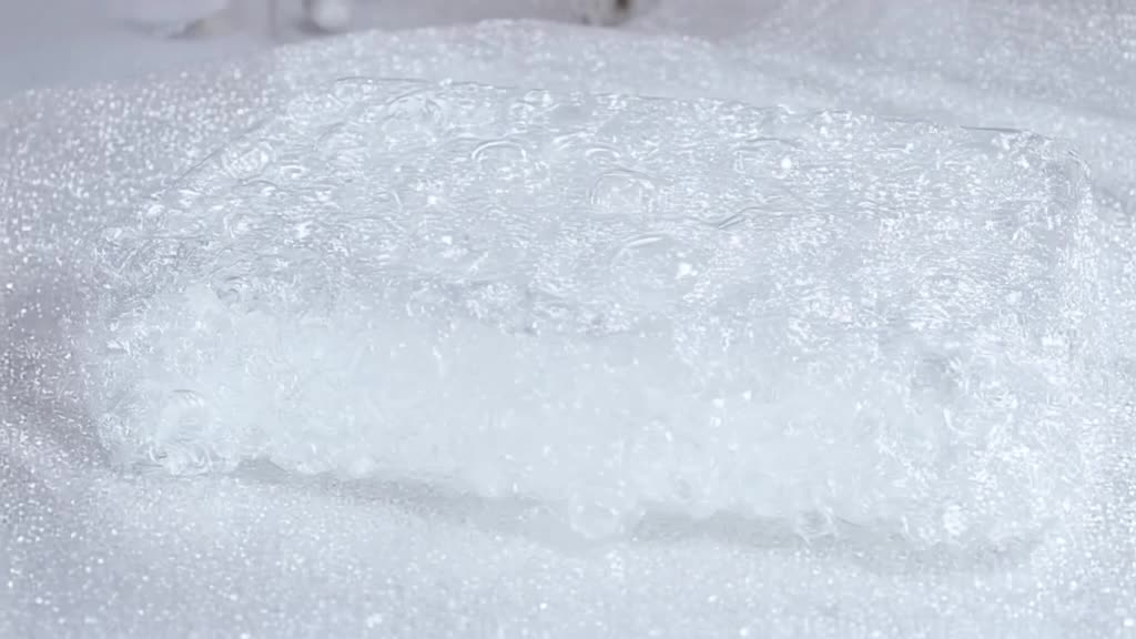 A slow-motion close-up of a bubble wrap sheet being pressed flat by a heavy glass block. …