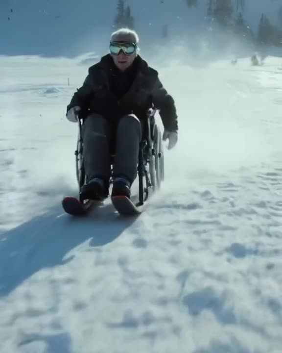 An elderly man in a wheelchair racing down a ski slope on a specially adapted sled. He we…