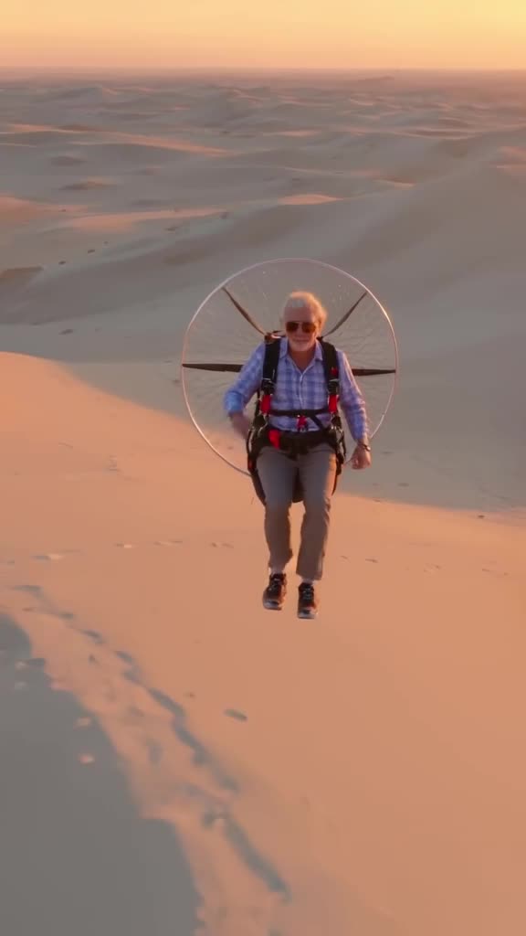 An old man in aviator shades paraglides over a pristine white desert dune. He tips his sh…