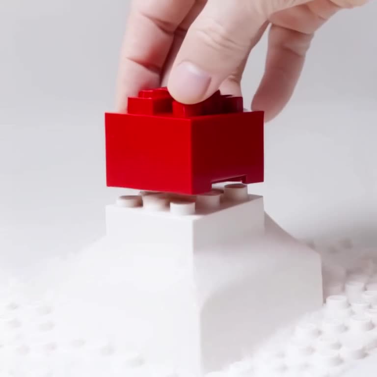 A robot hand delicately places a single red LEGO brick onto a mountain of white bricks. T…