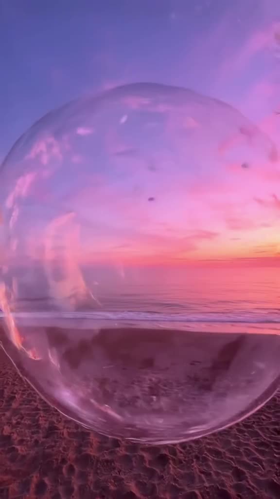 POV from inside a bubble machine on a beach at dusk. The camera is encased in a giant, sh…