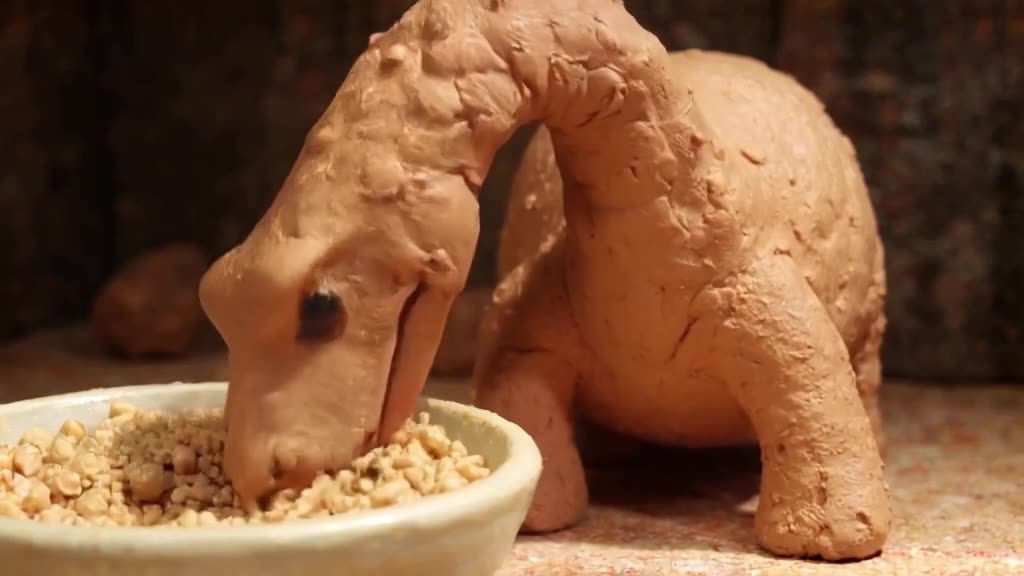 A claymation Brontosaurus stretches its long neck from a bowl of oatmeal. The neck elonga…