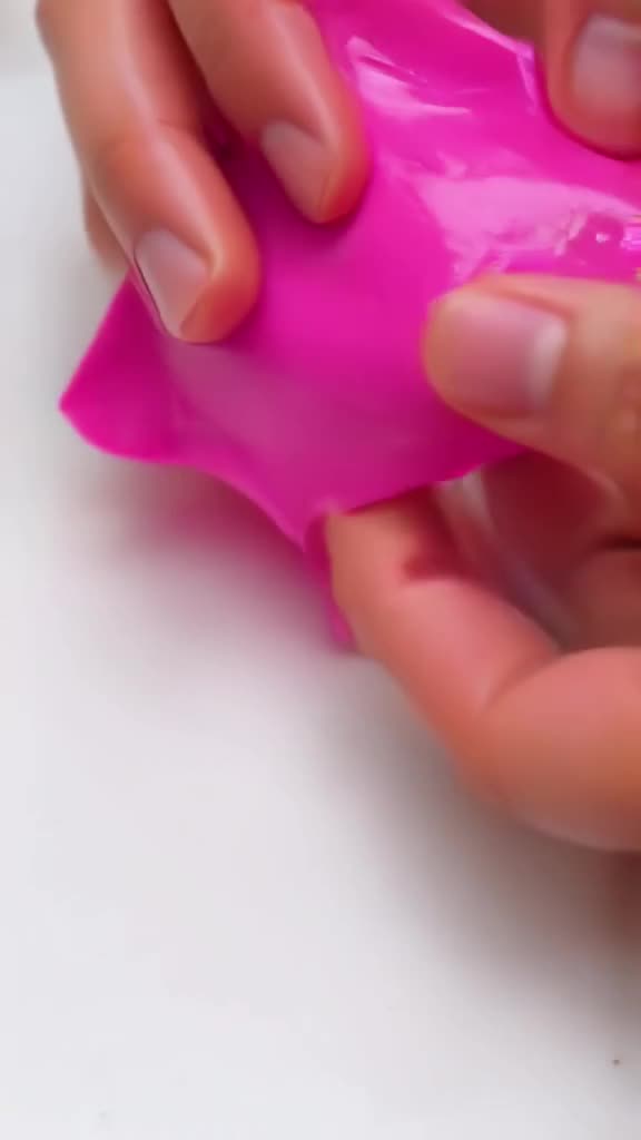Top-down view: tiny hands pour vibrant pink slip over a white tile. The liquid spreads be…