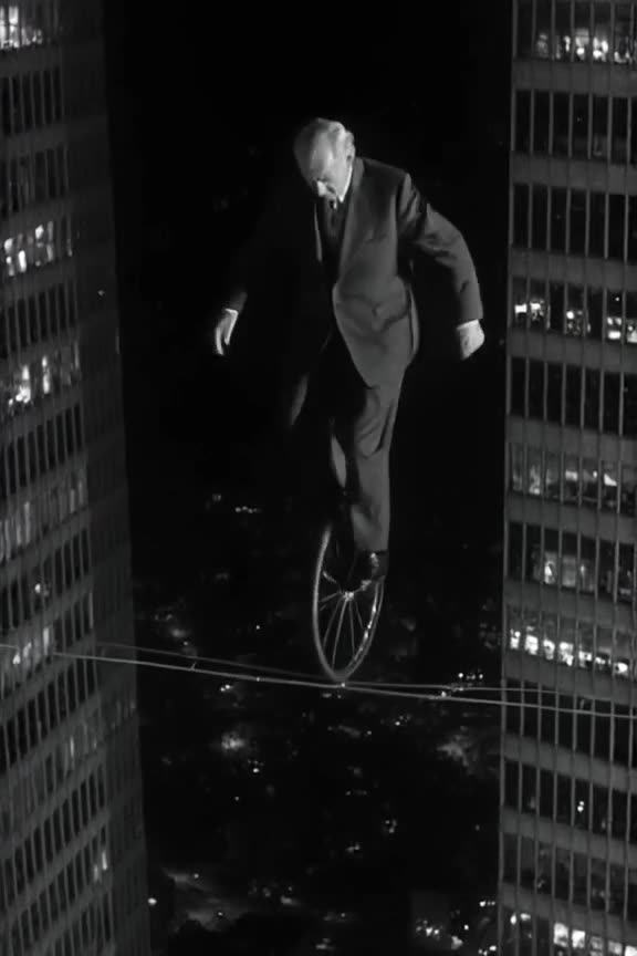 An elderly man in a suit rides a unicycle on a tightrope strung between two skyscrapers a…
