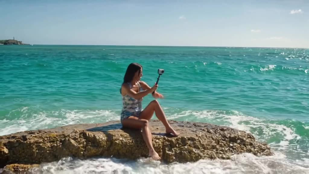 Medusa sits by the turquoise Aegean Sea, using a selfie stick with a GoPro. She adjusts h…