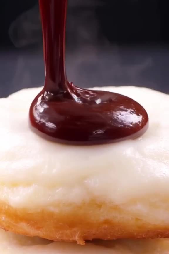 Thick, glossy chocolate syrup is poured in a perfect spiral over a stack of white donuts,…