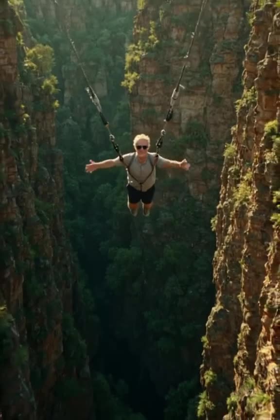 A senior citizen in sleek sunglasses bungee jumps from a bridge over a deep green canyon.…