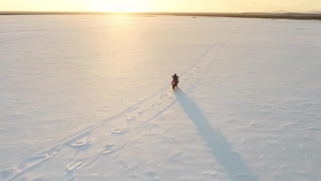 Drone shot of a motorcycle riding along a pristine white salt flat. The bike leaves track…