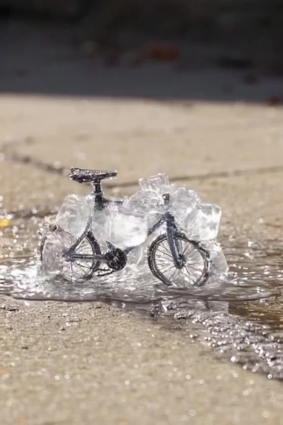 A tiny bicycle made of ice cubes melts slowly on a hot sidewalk, water droplets pooling a…