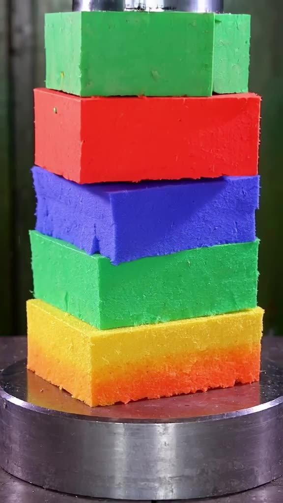 A hydraulic press crushes a stack of vibrant, multicolored foam shapes arranged in a perf…