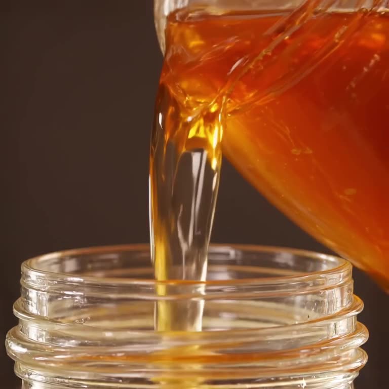 Golden syrup pours in slow motion into a glass jar, perfectly filling it without a single…