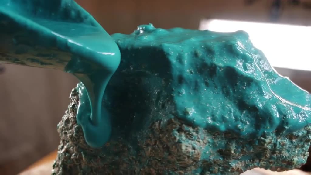 A hand pours thick teal glaze over a rough, unglazed sculpture. The liquid coats every cr…