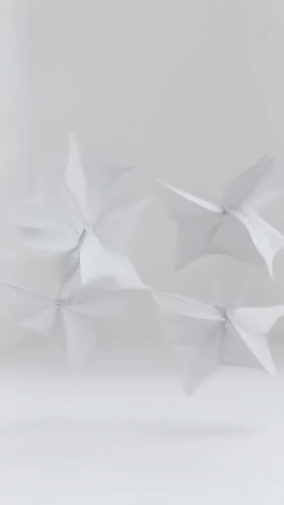 A horse made of folded paper origami flutters through a minimalist white room, each wing …