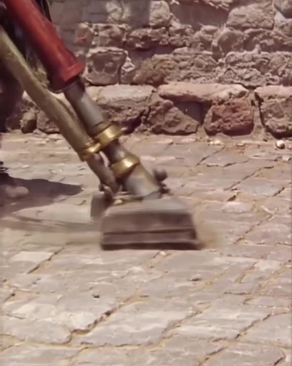 A medieval peasant uses a vacuum cleaner to clean a cobblestone street. The vacuum has a …
