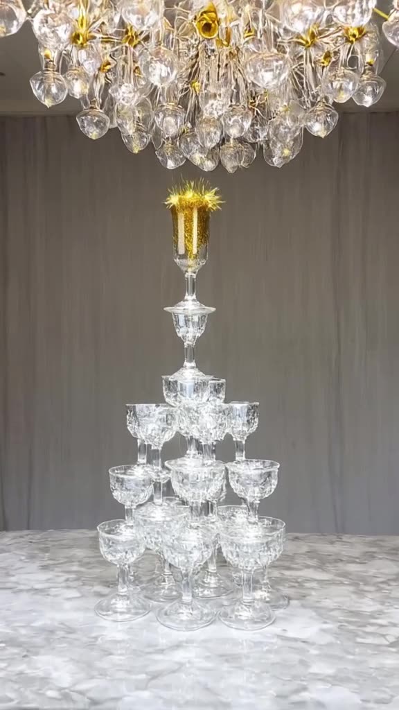 A tower of crystal champagne glasses stacks itself up by itself on a marble table. At the…
