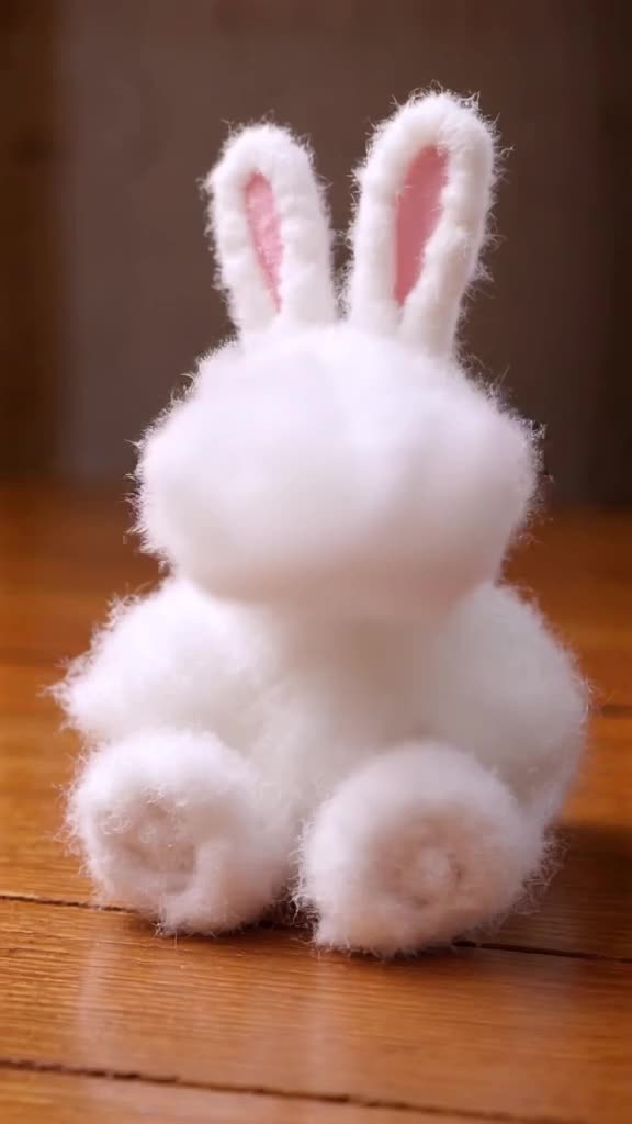 A fluffy white rabbit made of cotton candy sits on a wooden table. A gentle puff of breat…
