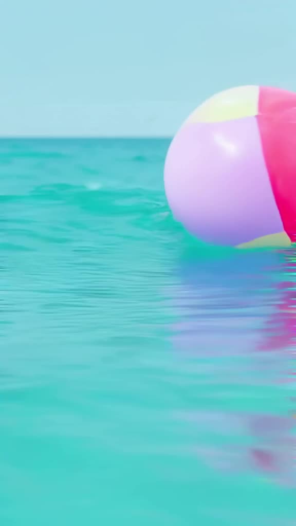 POV: A beach ball rolling into the gentle tide. Bright pastel colors against turquoise wa…