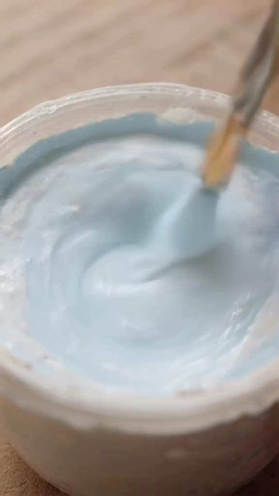 Paint mixing: a brush swirls white and light blue acrylic paint together in a jar. The co…