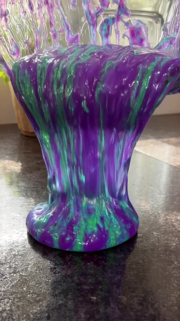 Thick, iridescent slime pours from a glass blender onto a dark countertop. It forms a per…