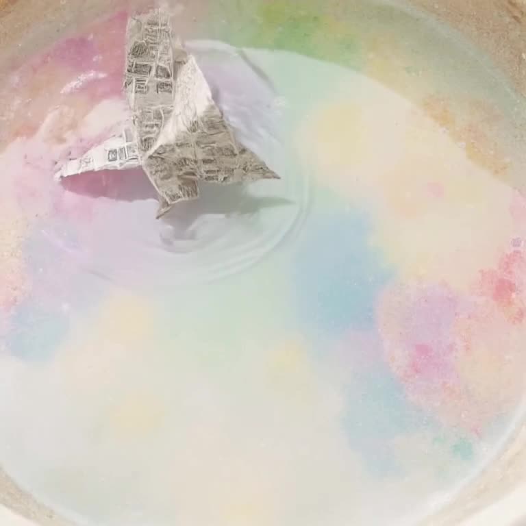 A origami crane folded from newspaper floating gently down into a tub of colorful soapy w…