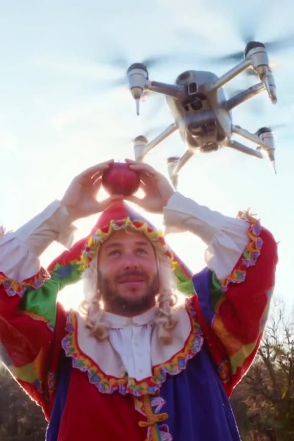 A medieval jester in motley colors tries to balance a red apple on his head while a moder…