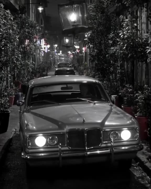 A silver vintage sedan tries to squeeze through a narrow alley lined with colorful potted…